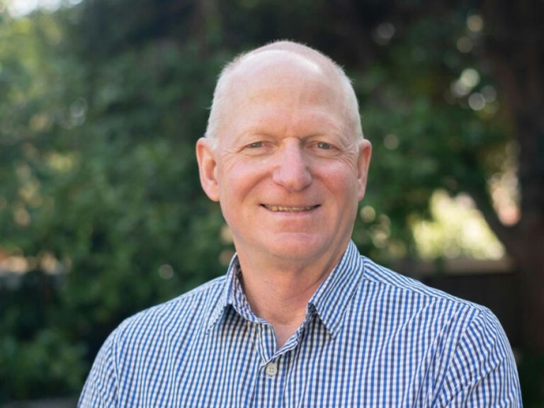 Tim has silver hair and is wearing a blue-checked shirt, while smiling at the camera.