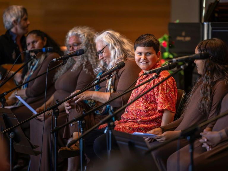 A First Nations Choir sit in a line, preparing to sing together. They have microphones.